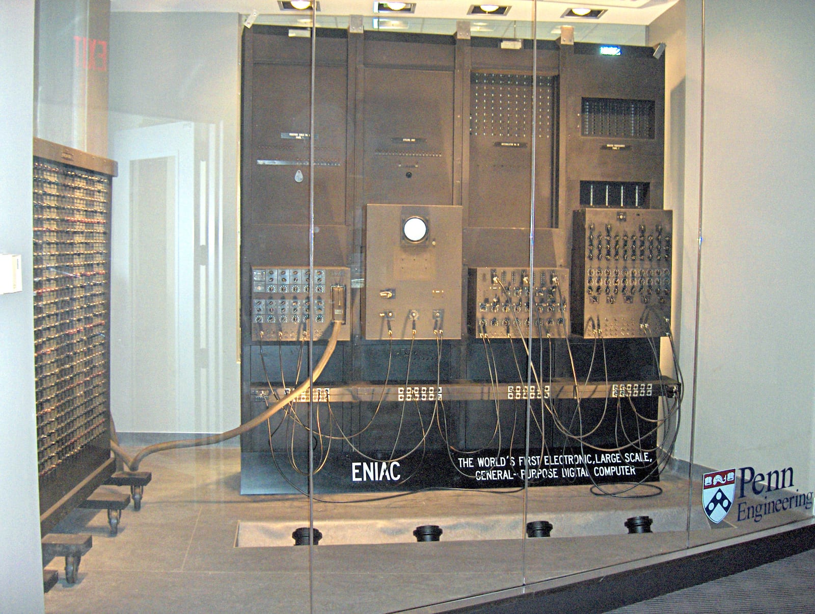 Two people standing beside the room-sized ENIAC computer covered in cables and switches